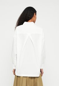 Camisa branca de manga comprida com um detalhe em painel nas costas, ajuste descontraído e gola. O tecido parece leve e suave.