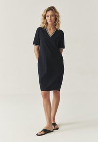 Robe à manches courtes bleu marine avec un col en V, bord blanc sur les manches, deux poches avant et une coupe décontractée. Portée avec des sandales noires.