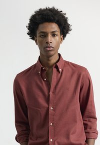 Camisa de botones en color óxido, hecha de algodón texturizado, con un cuello extendido, mangas largas arremangadas hasta el codo y un sutil detalle del logo.