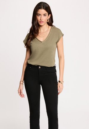 Femme aux longs cheveux bruns portant un haut vert olive à col en V, un pantalon noir, des boucles d'oreilles anneaux dorés, des colliers superposés et des bracelets.