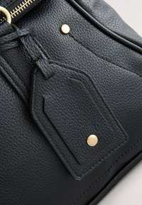 Sac en cuir noir texturé avec étiquette en cuir attachée, clous en métal doré et détail de fermeture éclair.