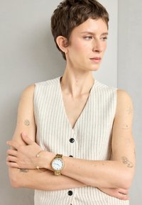 Gilet sans manches beige à rayures avec un décolleté en V profond, porté avec une montre dorée et un bracelet doré fin, mettant en valeur des tatouages sur les bras.