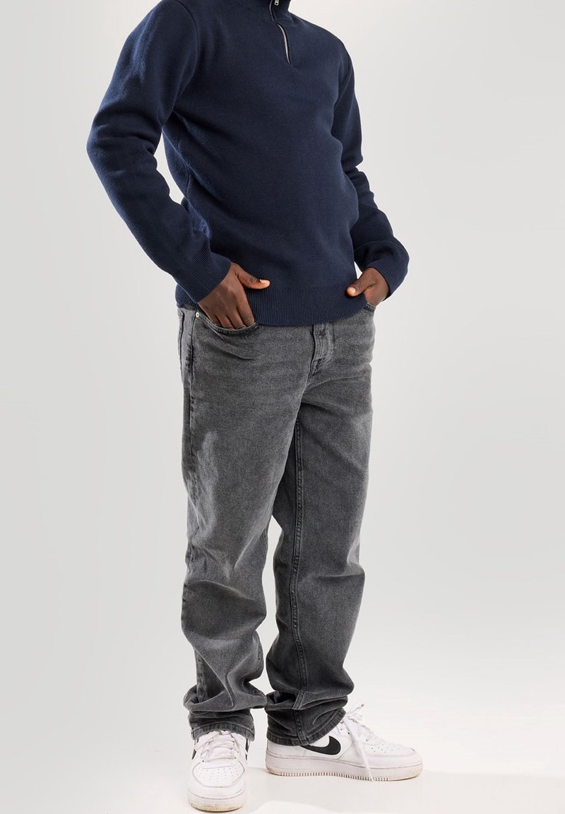 Pull à col zippé bleu marine avec manches longues, associé à un jean gris en denim et des baskets blanches avec un logo swoosh noir.