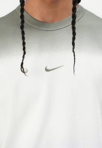 Lengvas medvilnės marškinėliai, turintys gradientą nuo alyvuogių žalios iki baltos, su mažuoju žaliu Nike logotipu ant krūtinės.