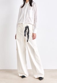 Vit randig skjorta med knäppning och en bröstficka, kombinerad med vida vita byxor och ett svart mönstrat slips. Svarta skor med stängd tå.