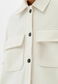 Cappotto con bottoni di colore crema chiaro, con colletto, due tasche sul petto, bottoni neri e un tessuto morbido e testurizzato.