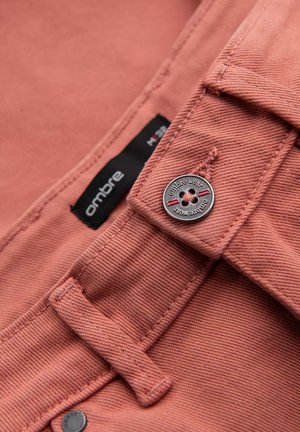 Chemise en coton couleur rose poussiéreux avec une finition texturée, dotée d'un bouton et d'une étiquette. Inclut une boucle pour le suspendre.