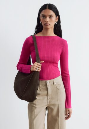 Jeune femme aux longs cheveux foncés portant un pull côtelé rose vif, un pantalon beige, des boucles d'oreilles créoles en argent et portant un grand sac à bandoulière marron foncé.