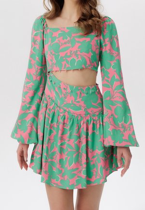 Kvinde iført et grøn- og pinkblomstret todelt sæt med lange klokkeærmer og udskæring i taljen mod en hvid baggrund.