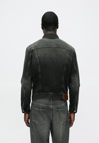 Čierna rifľová bunda s vybledlým efektom, manžetami na gombíky a hnedou koženou nášivkou na zadnej strane. Má klasický golier a dve zadné vrecká.