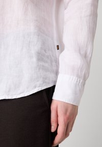 Camisa de lino blanca con un corte relajado, que presenta un puño rematado y una superficie texturada. Etiqueta visible en el lado. Pantalones oscuros parcialmente visibles.