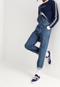 Pantalones de mezclilla con tirantes ajustables, corte holgado y puños enrollados, combinados con zapatillas de tablero de ajedrez moradas y calcetines blancos.