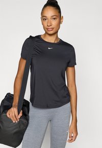 Μαύρο αθλητικό κοντομάνικο μπλουζάκι Nike με στενή εφαρμογή. Φορεμένο με γκρι κολάν, κρατώντας μια μαύρη τσάντα γυμναστηρίου. Έμφαση σε απλό, λειτουργικό σχεδιασμό.