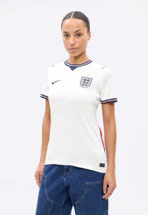 Frau trägt weißes England-Fußballtrikot mit marineblauen und roten Akzenten, kombiniert mit blauer Jeanshose, steht vor einfachem Hintergrund.