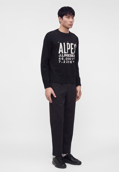 Jeune homme debout portant un pull noir avec un texte blanc, un pantalon noir, des chaussettes blanches et des baskets noires sur un fond clair uni.