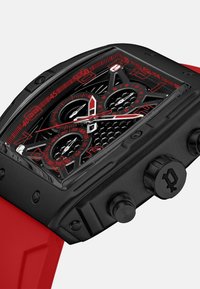 Juodas laikrodis su stačiakampiu korpusu, raudonomis detalėmis, raštuotu ciferblatu, dviem subciferblatais ir tekstūruota guma dirželiu. Yra apvalių mygtukų ir karūna.