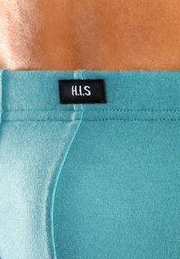 Mėlynos spalvos medvilniniai apatiniai su lygiu tekstūru, su juodu etiketu, kuriame siuvinėjama "H.I.S" ant juosmens. Artimas vaizdas, akcentuojantis siūles.