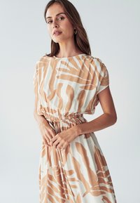 Lehká krémová šaty s hnědými abstraktními pruhy, krátké rukávy a nařasený pas. Obsahuje šňůrku pro nastavitelnou velikost.