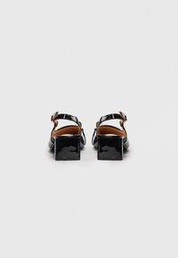 Zwarte patenten leren lage blokhak sandalen met enkelbandjes, van achteren weergegeven op een effen witte achtergrond.