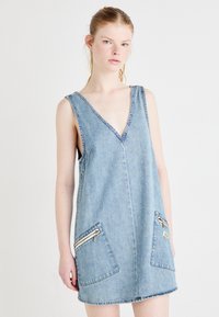 Denim ilman hihoja oleva mekko, jossa on syvä V-pääntie ja kaksi etutaskua vetoketjulla. Vaalea sininen väri, arkinen muotoilu ja hapsutettu helma.