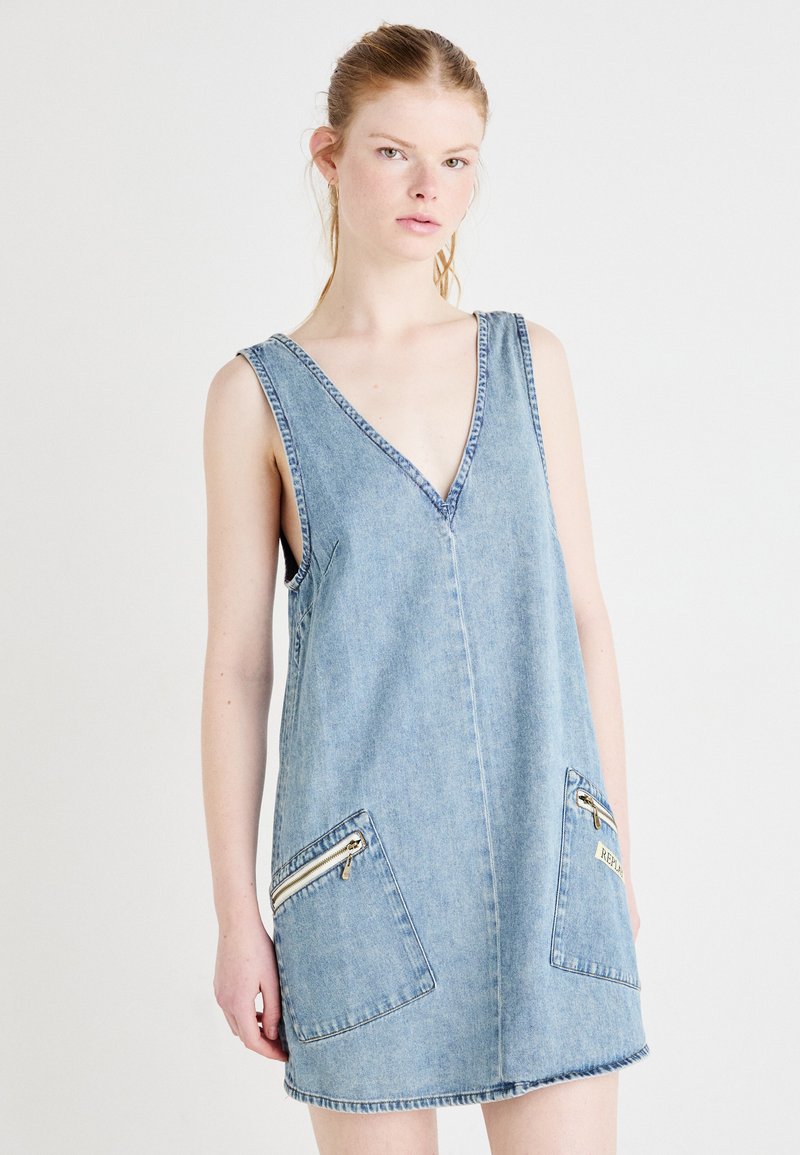 Denim ilman hihoja oleva mekko, jossa on syvä V-pääntie ja kaksi etutaskua vetoketjulla. Vaalea sininen väri, arkinen muotoilu ja hapsutettu helma.