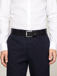 Cinturón de cuero negro con hebilla plateada, usado sobre pantalones oscuros de color marino. Presenta un pequeño detalle de logo en el cinturón. Textura suave.
