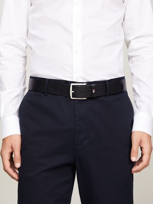 Ceinture en cuir noir avec une boucle en argent, portée sur un pantalon bleu marine foncé. Présente un petit logo sur la ceinture. Texture lisse.