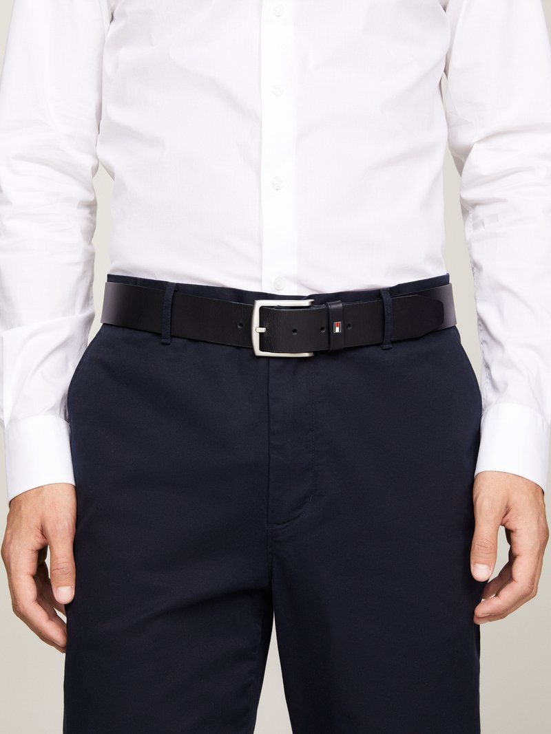 Cinturón de cuero negro con hebilla plateada, usado sobre pantalones oscuros de color marino. Presenta un pequeño detalle de logo en el cinturón. Textura suave.