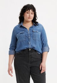 Camicia di jeans azzurro chiaro con bottoni e due tasche sul petto, maniche arrotolate, indossata su jeans neri a vita alta con una finitura testurizzata.
