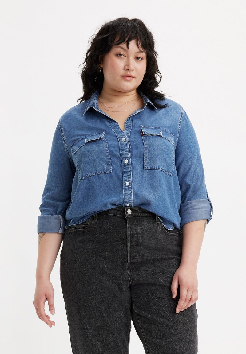 Camicia di jeans azzurro chiaro con bottoni e due tasche sul petto, maniche arrotolate, indossata su jeans neri a vita alta con una finitura testurizzata.