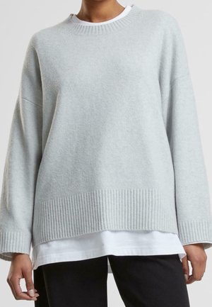 Personne portant un pull tricoté ample gris clair par-dessus une chemise blanche et un pantalon noir, visible des épaules aux hanches.