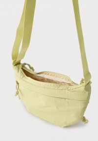 Ελαφριά κίτρινη τσάντα χιαστί με ρυθμιζόμενο λουρί και επάνω φερμουάρ, ημικατεβασμένο για να φαίνεται το εσωτερικό από μπεζ ύφασμα.