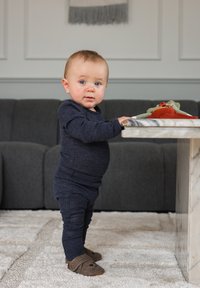 Temno modro bodi z dolgimi rokavi in ujemajoče hlače na malčku. Rjavi copati z živalskim vzorcem. Bela miza iz marmorja s pisanimi tkaninskimi igračami.