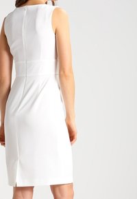 Weißes ärmelloses Kleid mit figurgerechter Passform, verdecktem Reißverschluss hinten, knielanger Schnitt und einem Schlitz in der Mitte hinten für Bewegungsfreiheit.