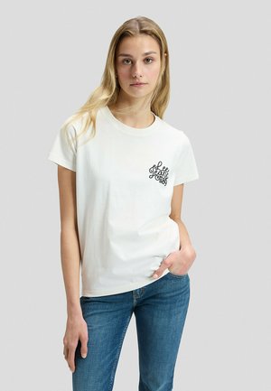 Młoda kobieta z długimi blond włosami stoi w białej koszulce z napisem ""Lee Jeans 1889"" i niebieskich dżinsach, z ręką w kieszeni, na jednolitym tle.