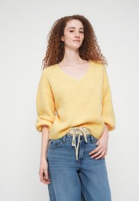 Νεαρή γυναίκα με κατσαρά κόκκινα μαλλιά, που φοράει ένα χαλαρό κίτρινο πλεκτό πουλόβερ και μπλε τζιν, στέκεται με το ένα χέρι στην τσέπη μπροστά σε λευκό φόντο.