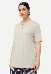 Top corto beige chiaro a maniche corte con scollatura a V e vestibilità rilassata. Tessuto morbido e liscio con un design minimalista. Abbinato a pantaloni blu scuro con motivo.