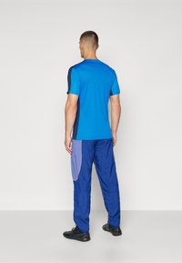 Blaues Sport-T-Shirt mit einem kontrastierenden dunkelblauen Seitenpaneel, kombiniert mit blauen Hosen, die hellere blaue Seitentaschen und orangefarbene Nähte aufweisen.