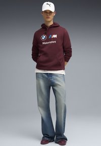 Felpa bordeaux con logo BMW Motorsport, t-shirt bianca sottostante, jeans azzurri chiari strappati e scarpe bordeaux. Cappellino bianco.