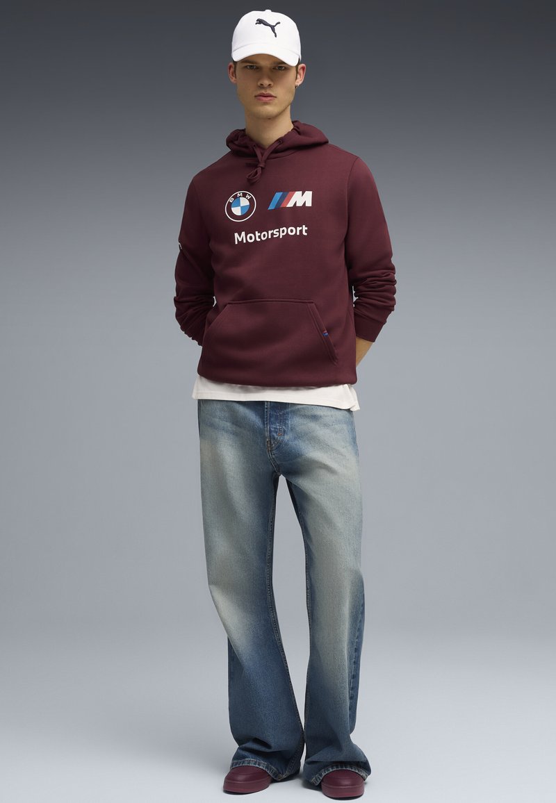 Felpa bordeaux con logo BMW Motorsport, t-shirt bianca sottostante, jeans azzurri chiari strappati e scarpe bordeaux. Cappellino bianco.