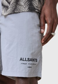 Pantaloni scurți gri deschis, realizați dintr-un material ușor, cu imprimeu negru pe piciorul stâng și o buzunar pe laterala.