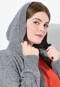 Grå stickad hoodie med en texturerad finish, som har en snörning i huvan och långa ärmar. Under den syns en enfärgad röd skjorta.