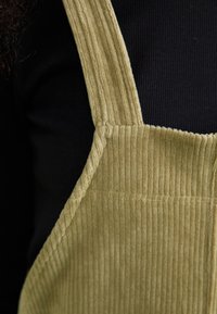 Le salopette in velluto a coste verde oliva presentano bretelle larghe e un motivo a coste testurizzato, abbinate a una maglietta a maniche lunghe nera sotto.