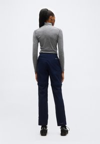 Γυναίκα με αλογοουρά που φοράει γκρι μπλουζάκι με μακριά μανίκια και ανοιγόμενο παντελόνι navy μπλε για εξωτερικούς χώρους, στέκεται γυρισμένη με την πλάτη σε απλό λευκό φόντο.