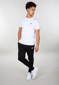 Ung mand står med hænderne i lommerne iført hvid t-shirt, sorte joggere med logos og hvide sneakers foran en ensfarvet baggrund.