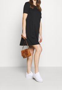 Donna in un vestito nero che tiene una piccola borsa marrone, indossando sneaker bianche, in piedi contro uno sfondo chiaro e uniforme.