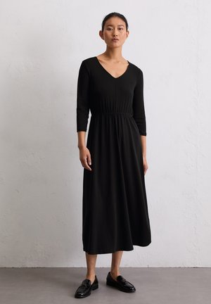 Zwarte lange mouw jurk met een V-hals, elastische taille en vloeiende silhouette; gestyled met zwarte loafers en geplaatst tegen een minimalistische achtergrond.