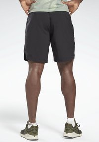 Vista trasera de una persona que lleva pantalones cortos deportivos negros, zapatillas verdes de Reebok, calcetines blancos y una camiseta ligera verde con puntos, con las manos en las caderas.