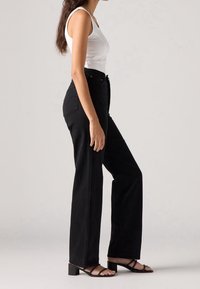 Femme debout de profil portant un haut blanc sans manches, un pantalon noir taille haute à jambes larges et des sandales noires ouvertes à talons blocs.