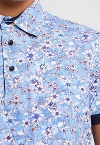 Ljusblå kortärmad skjorta med blommönster av vita och lila blommor. Knapparna är mörkblå. Tyget verkar ha en texturerad yta.